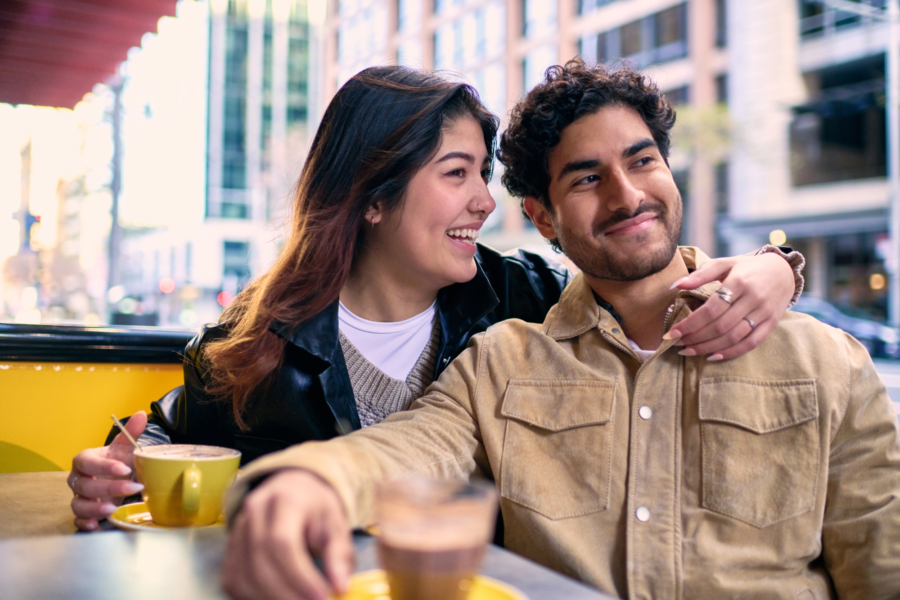 An image of a young couple smiling, sitting outdoors on a coffee shop patio